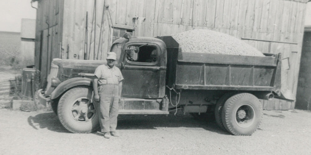 black and white image of a middle-aged man standing in front of a dump truck filled with small stones